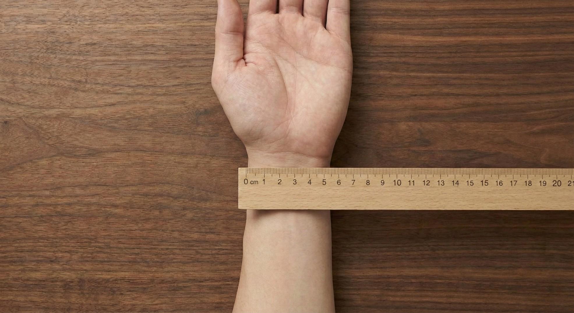 A wooden ruler being placed across the top of a wrist to measure its flat width bone to bone.