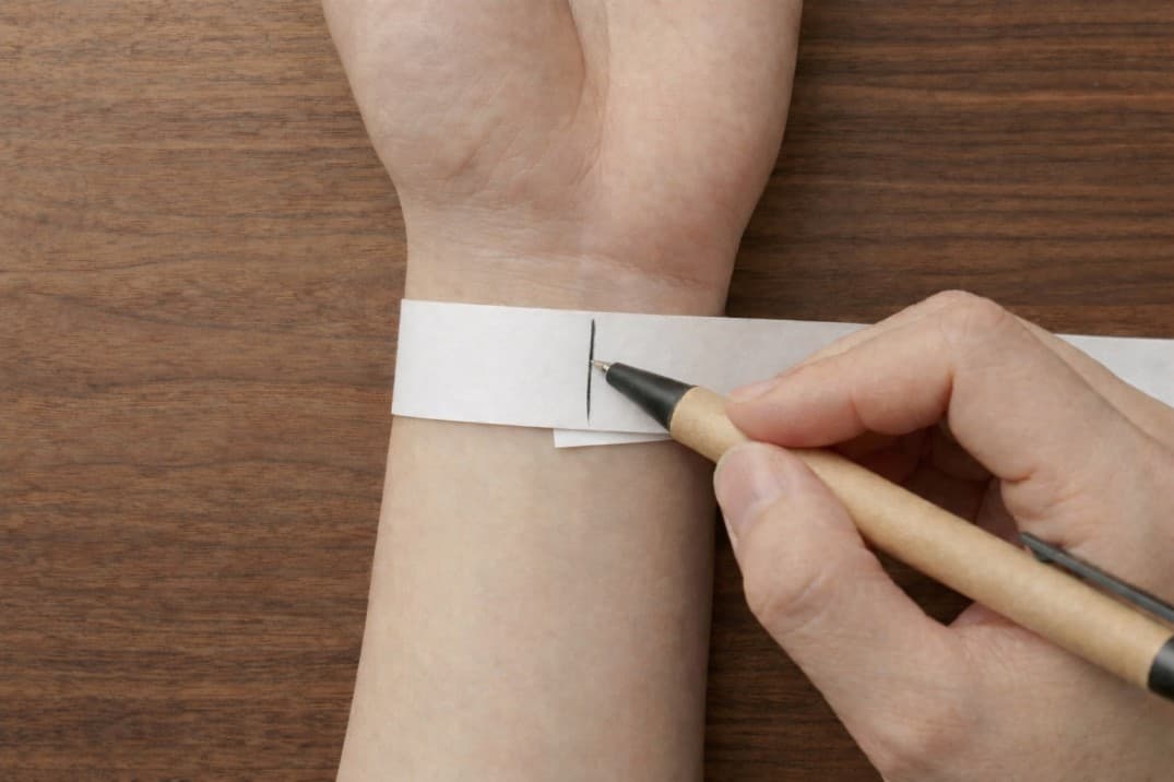 Marking the exact overlap point on a paper strip with a pen to find the wrist circumference. Marking the exact overlap point on a paper strip with a pen to find the wrist circumference.